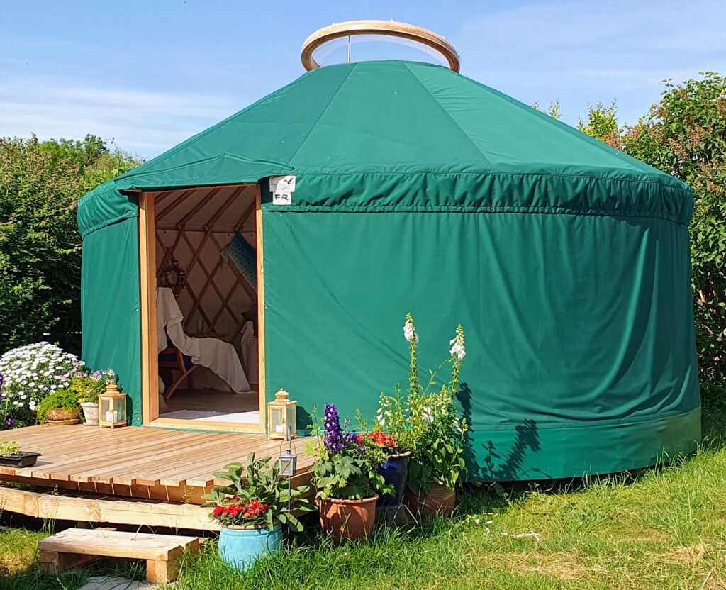 Jurten set udefra på en solskinsdag med blå himmel. Foran døren er en terrasse, hvor flere store krukker fyldt med farverige blomster står i fuldt flor.